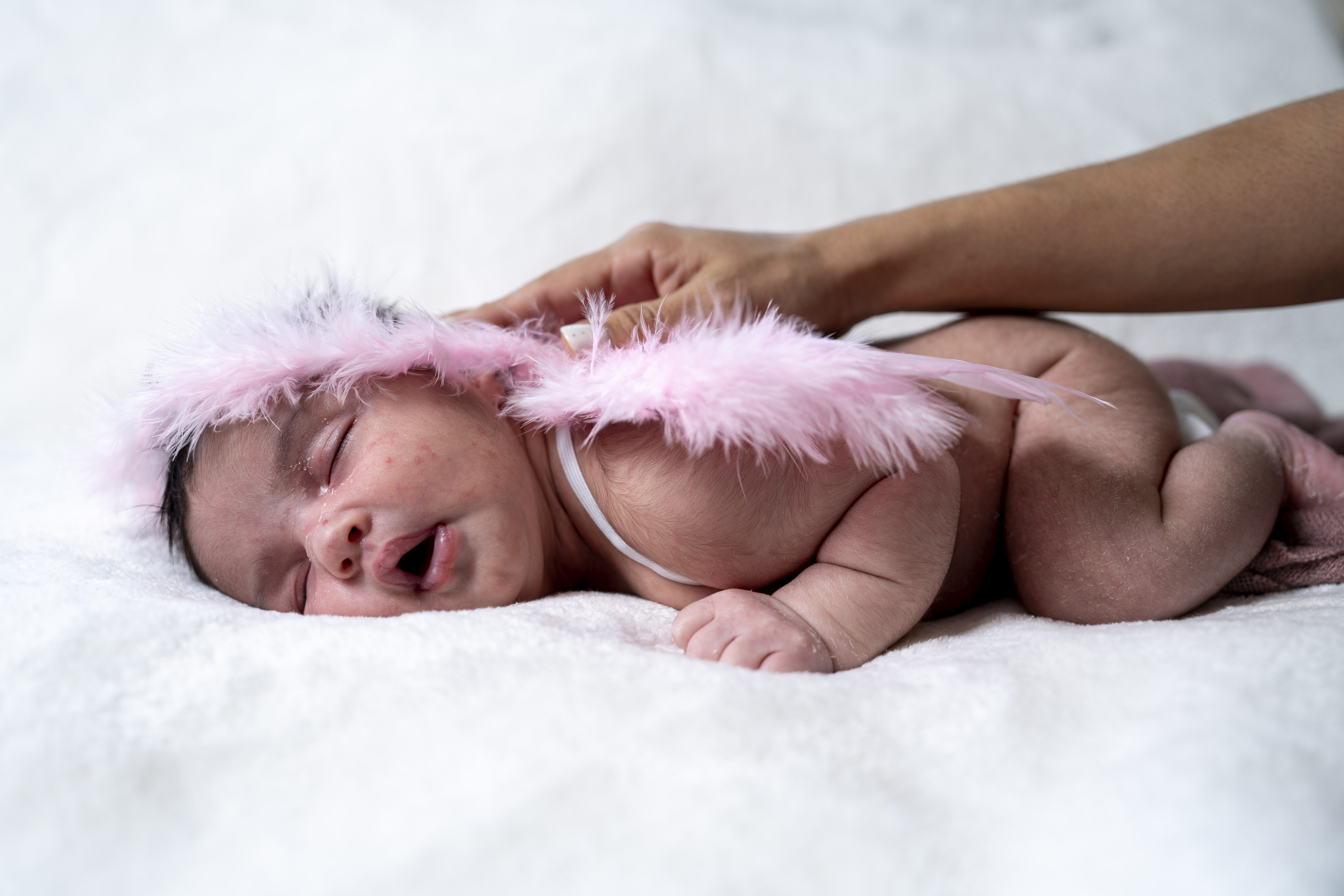 A baby wearing a pink feathered headband, lying on a white blanket.