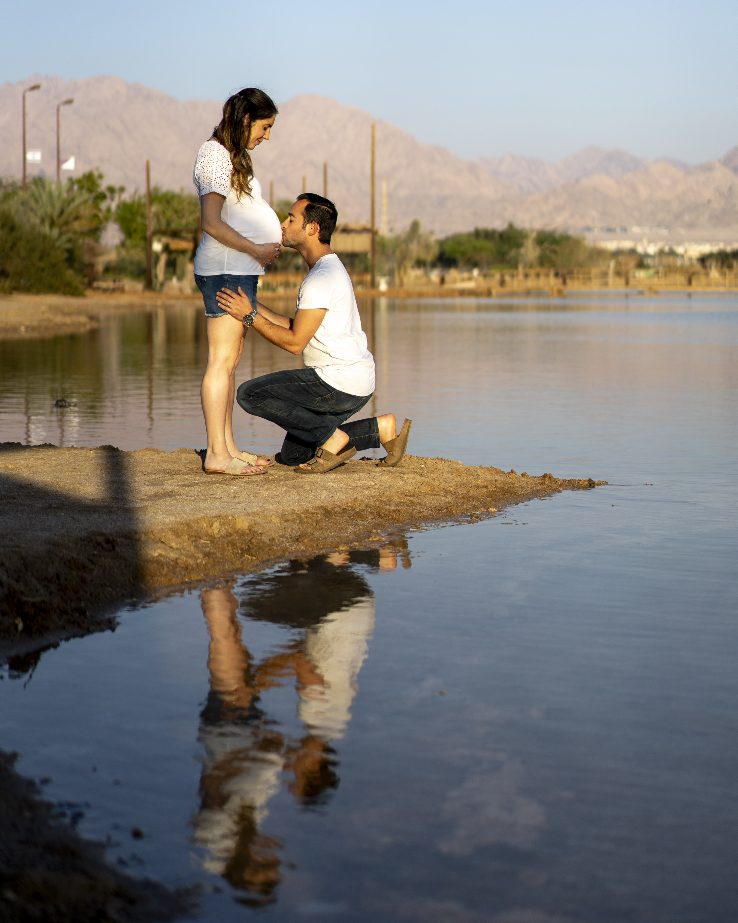 A man and woman, both wearing white t-shirts, stand on a rocky outcropping in a body of water, with a mountain range in the background.