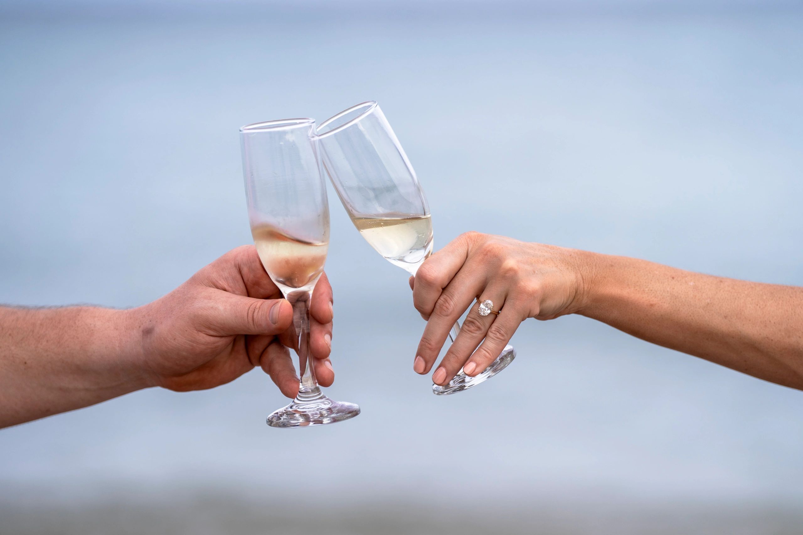 A man and woman's hands clinking champagne flutes together, with the woman's left hand showing an engagement ring.