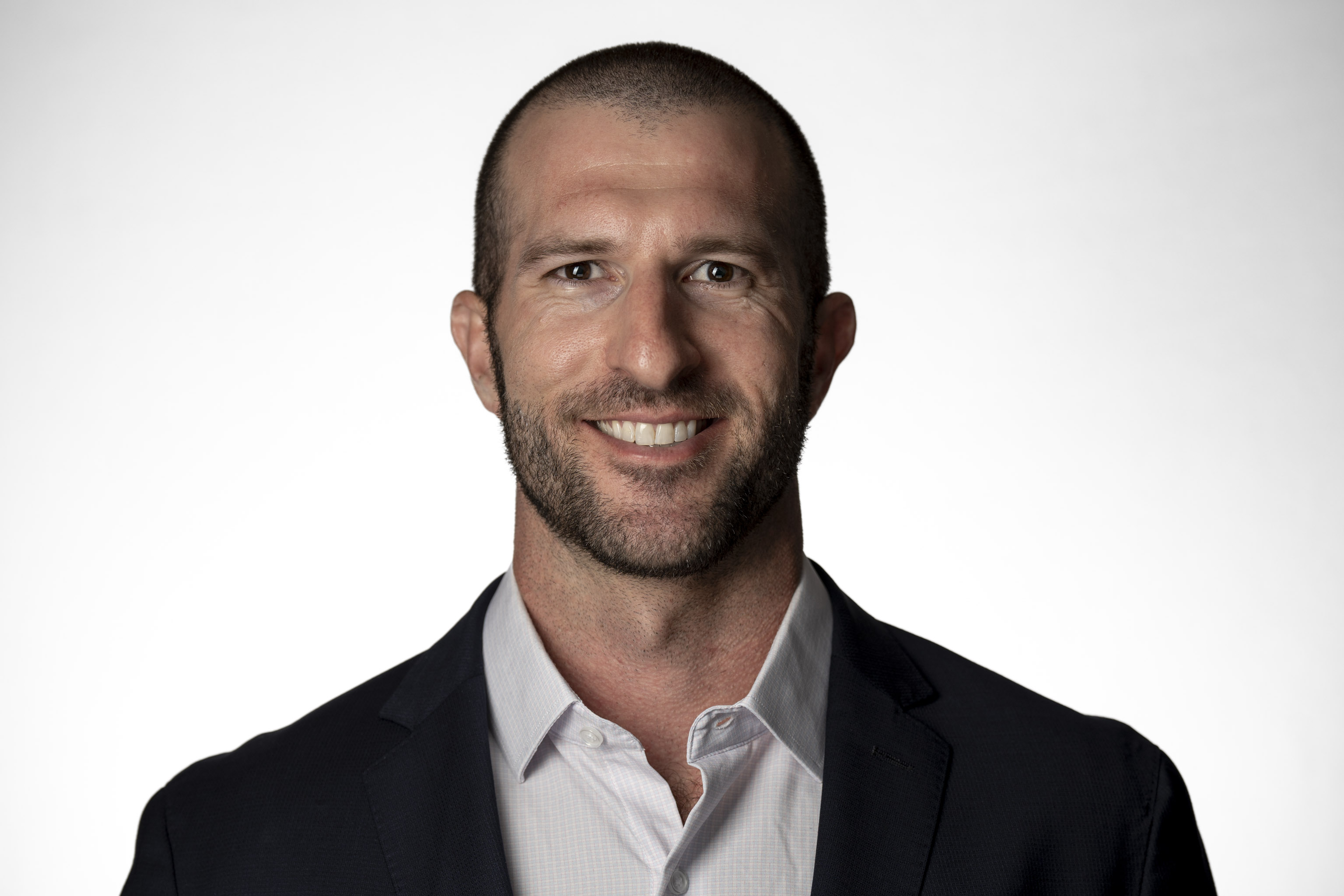 Man in a dark suit jacket and white collared shirt, smiling against a neutral grey backdrop.