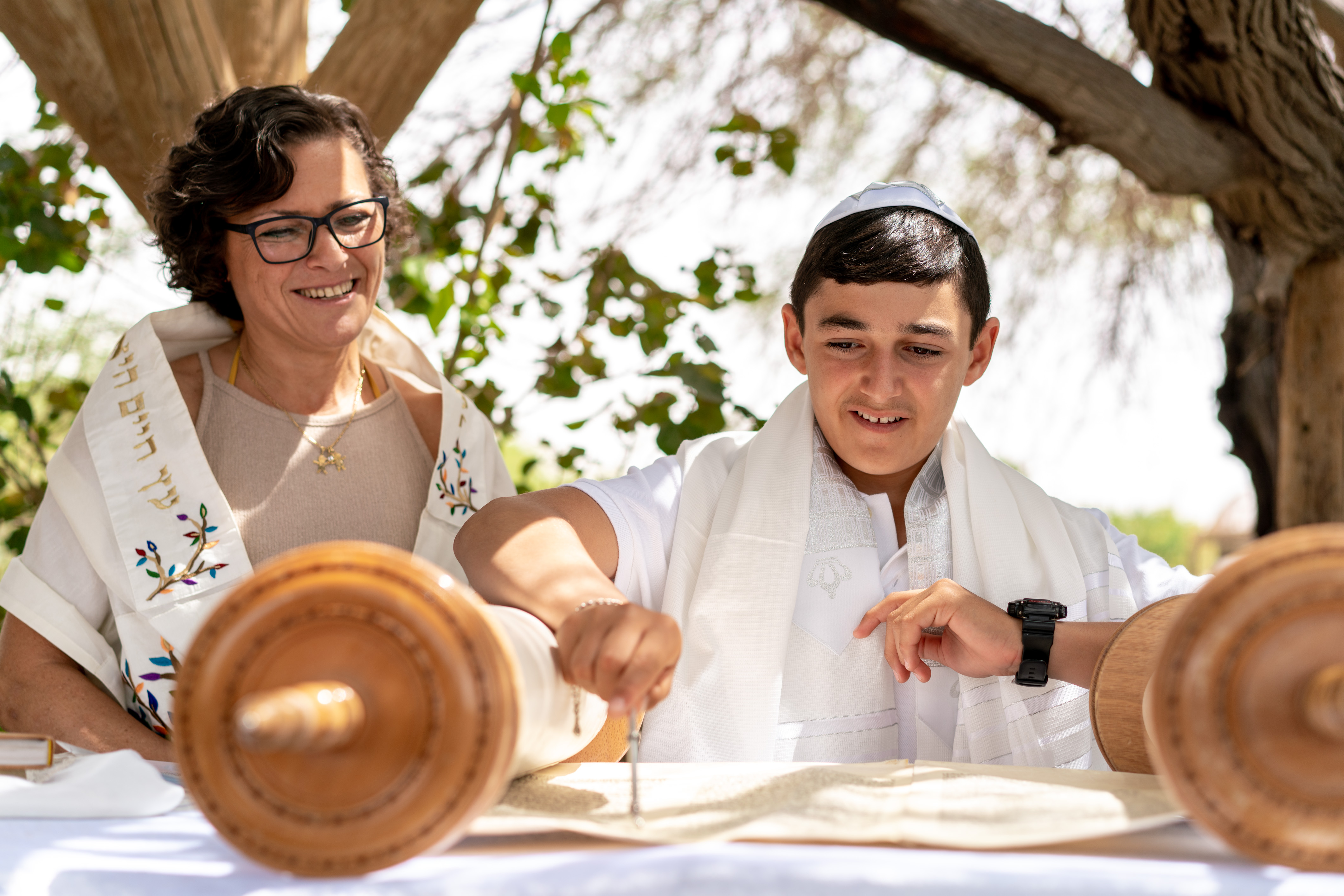 A boy in a white shirt and kippah, smiling at the camera, with a woman in a white dress and shawl standing next to him.