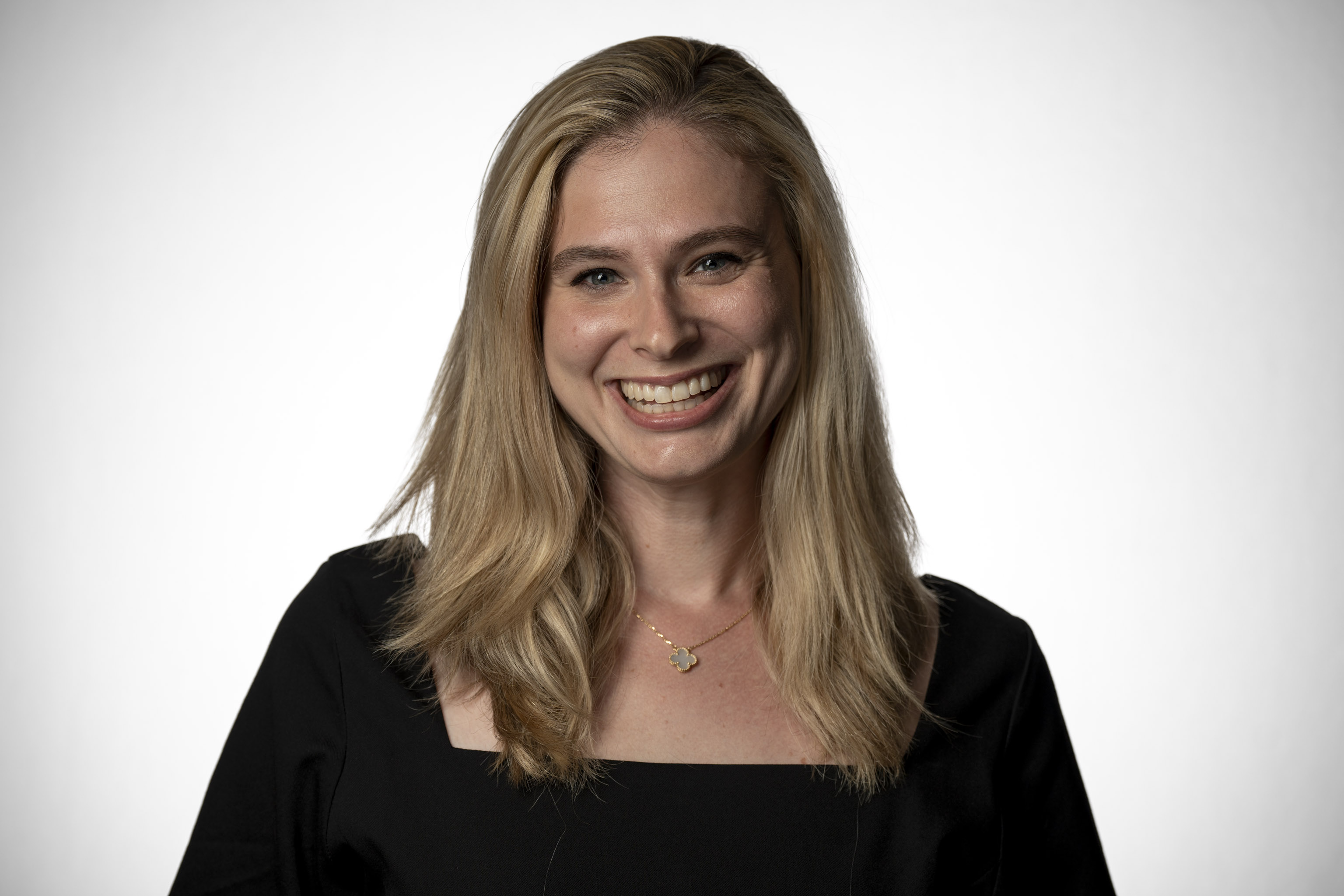 A woman with long blonde hair and a black top smiles at the camera against a white backdrop.
