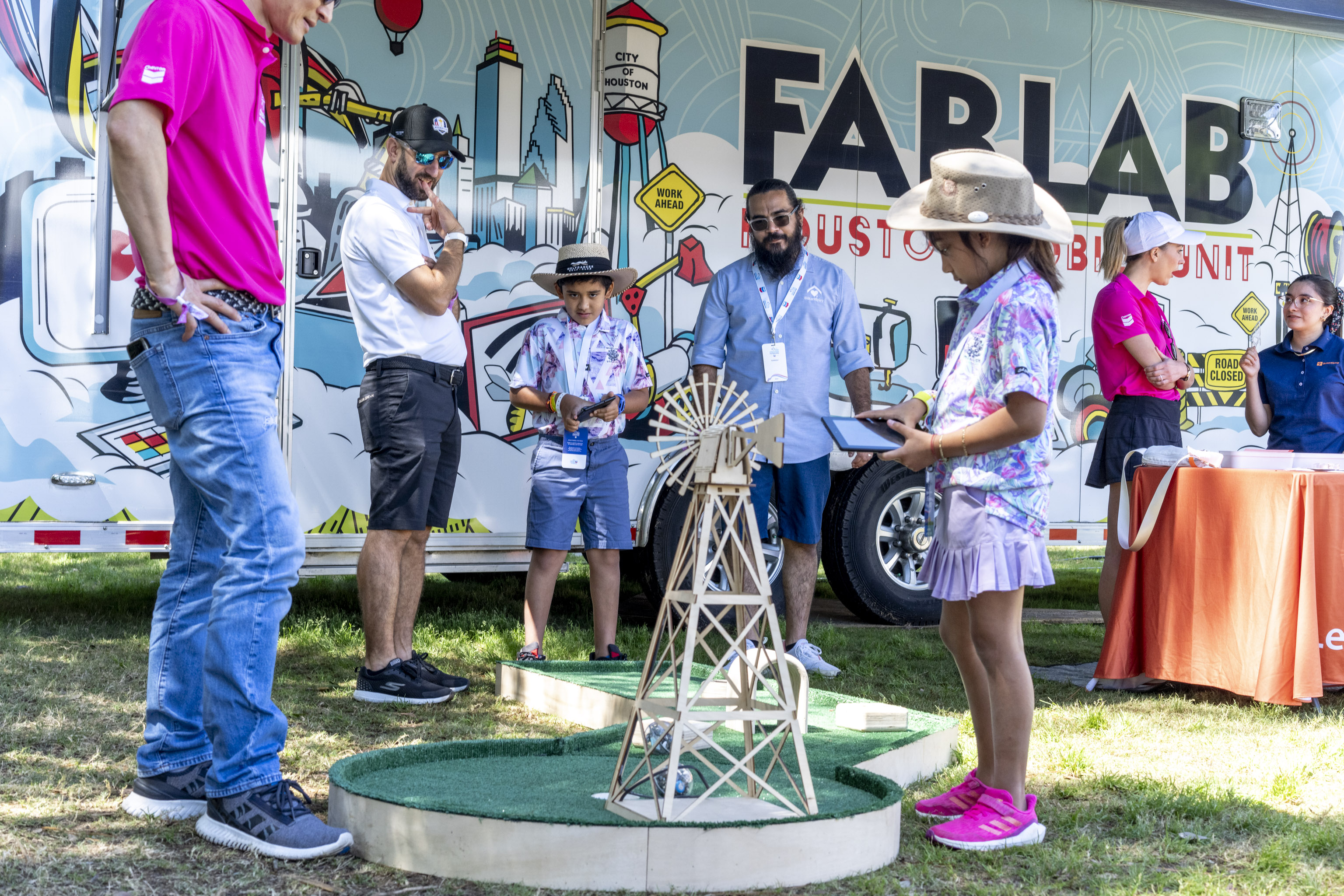A young girl in a floral shirt and purple skirt stands in front of a miniature golf course, holding a tablet and looking at a model windmill.