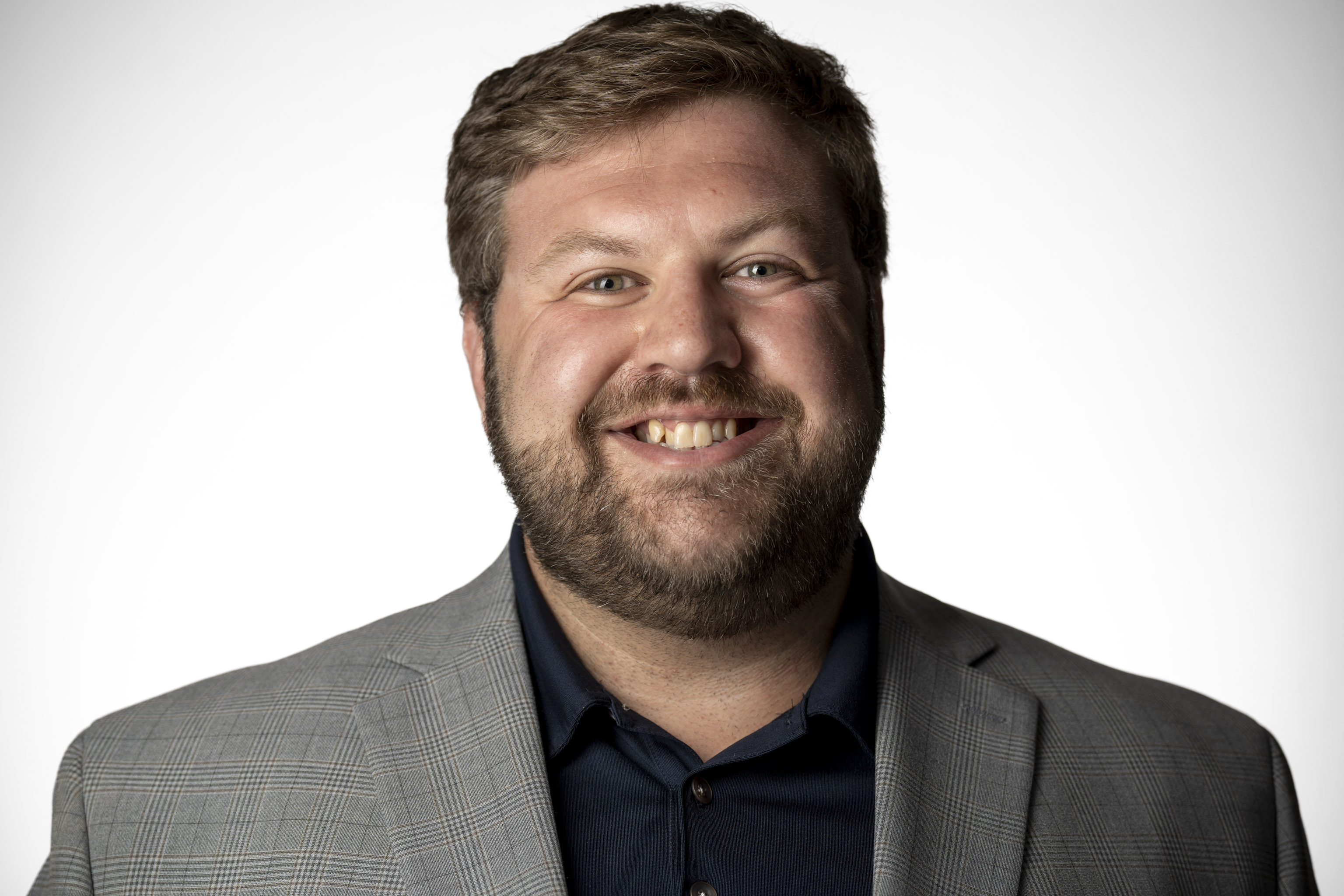 Man in a gray plaid suit jacket and dark blue collared shirt, visible from the chest up, against a plain white backdrop.