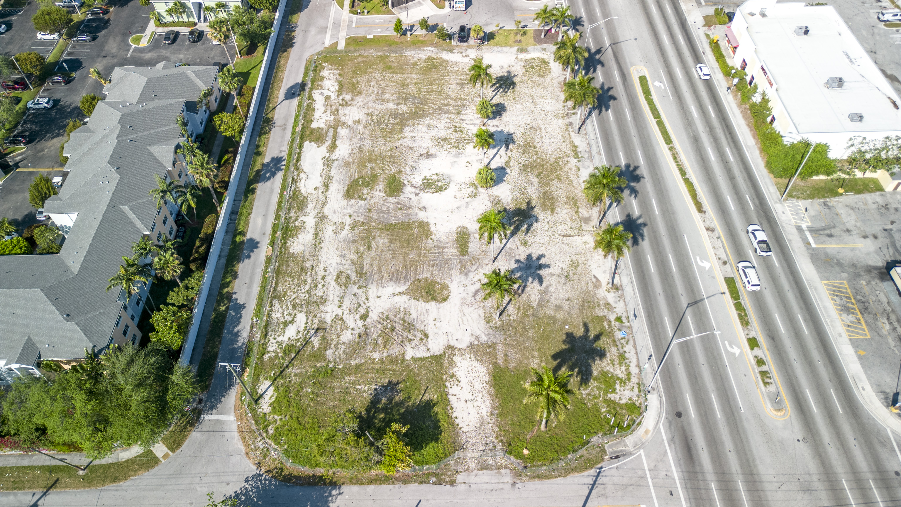 Aerial view of a vacant lot in a residential area, surrounded by palm trees and a road.